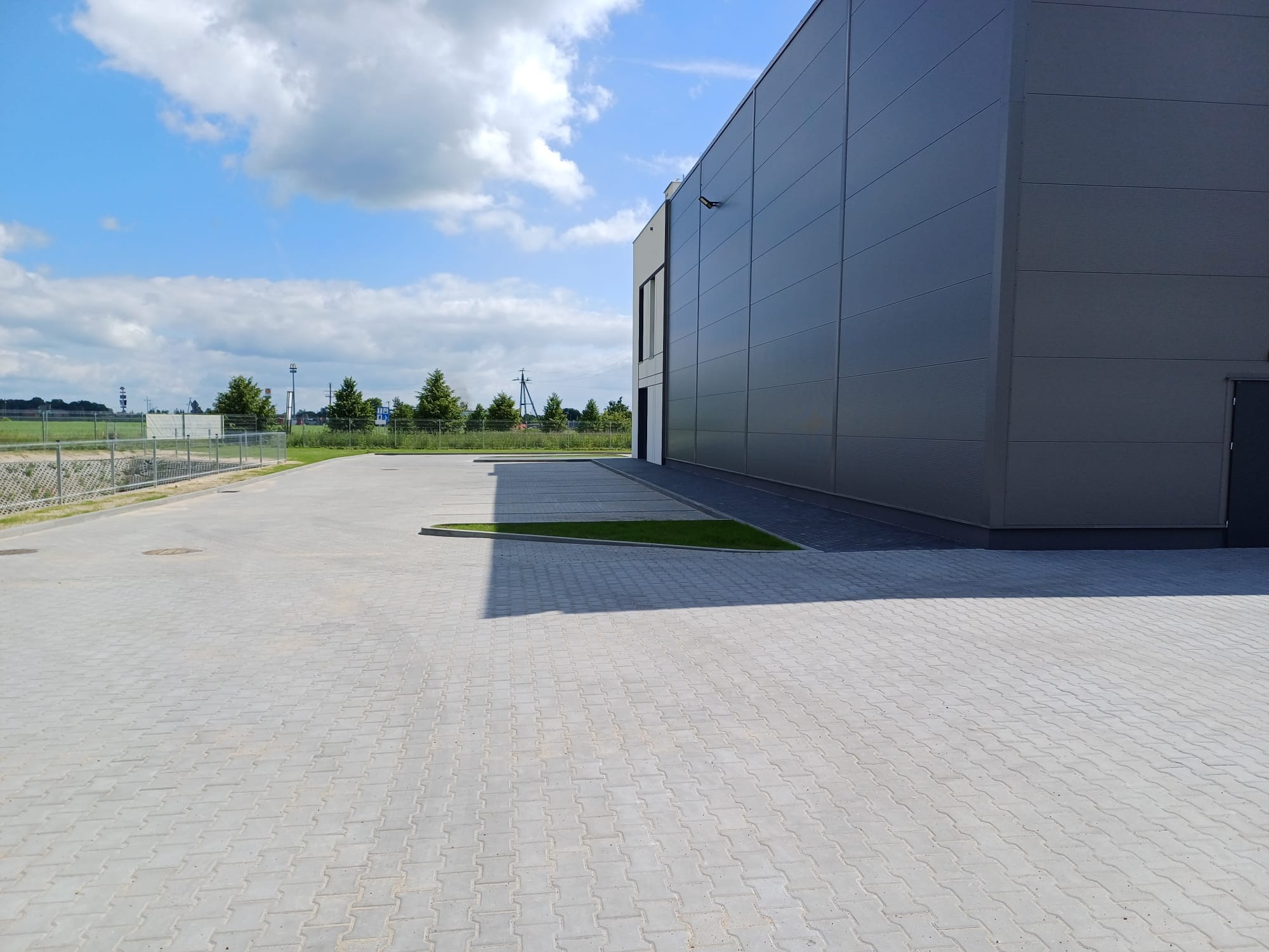 Szara kostka brukowa ułożona przed nowoczesnym budynkiem przemysłowym w słoneczny dzień. Widoczne trawniki i ogrodzenie w tle.