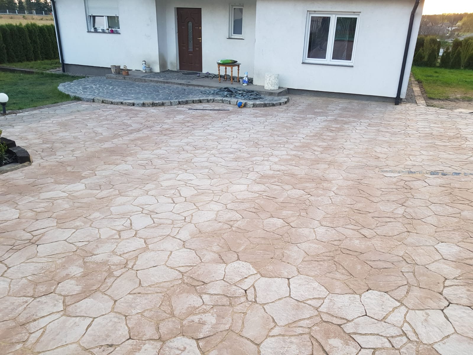 Wykończony podjazd z nieregularną, beżową nawierzchnią imitującą kamień, prowadzący do białego domu z brązowymi drzwiami i oknami. Widoczne elementy dekoracyjne.