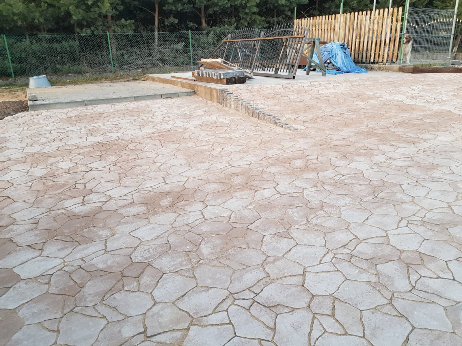 Ukończona nawierzchnia z nieregularnej kostki betonowej w odcieniach beżu i szarości, z widocznym fragmentem obrzeża i przygotowanym podjazdem. W tle ogrodzenie i zieleń.