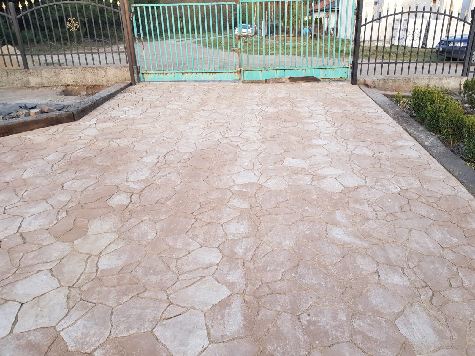Ułożona nawierzchnia z nieregularnych płyt kamiennych w odcieniach beżu, prowadząca do metalowej bramy wjazdowej. Widoczne krzewy i fragmenty niezakończonych prac.
