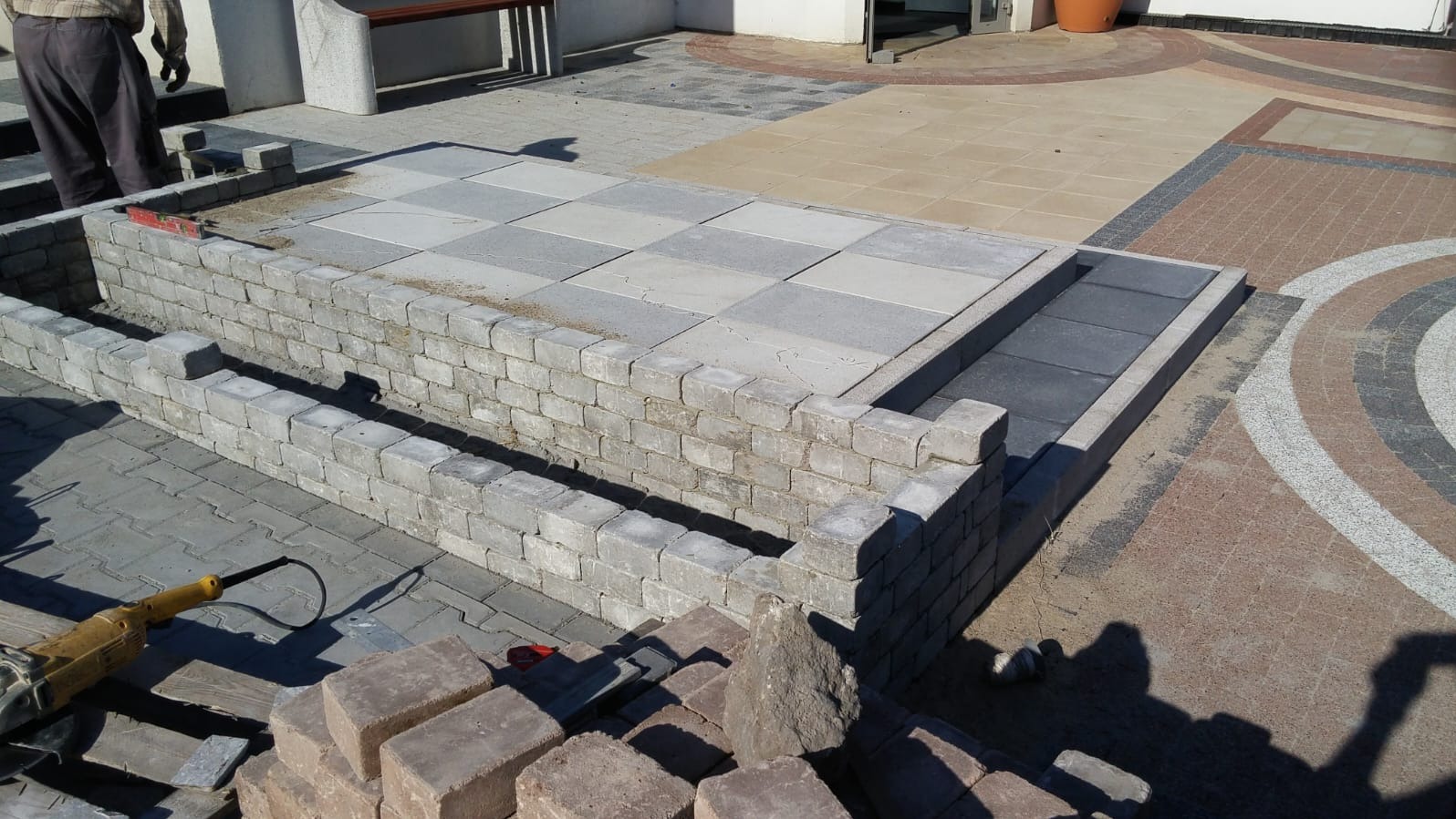 Budowa murku z kostki, widoczne narzędzia i materiały. Częściowo ułożona nawierzchnia z kostki w szachownicę. Prace brukarskie w toku, detale wykończeniowe.