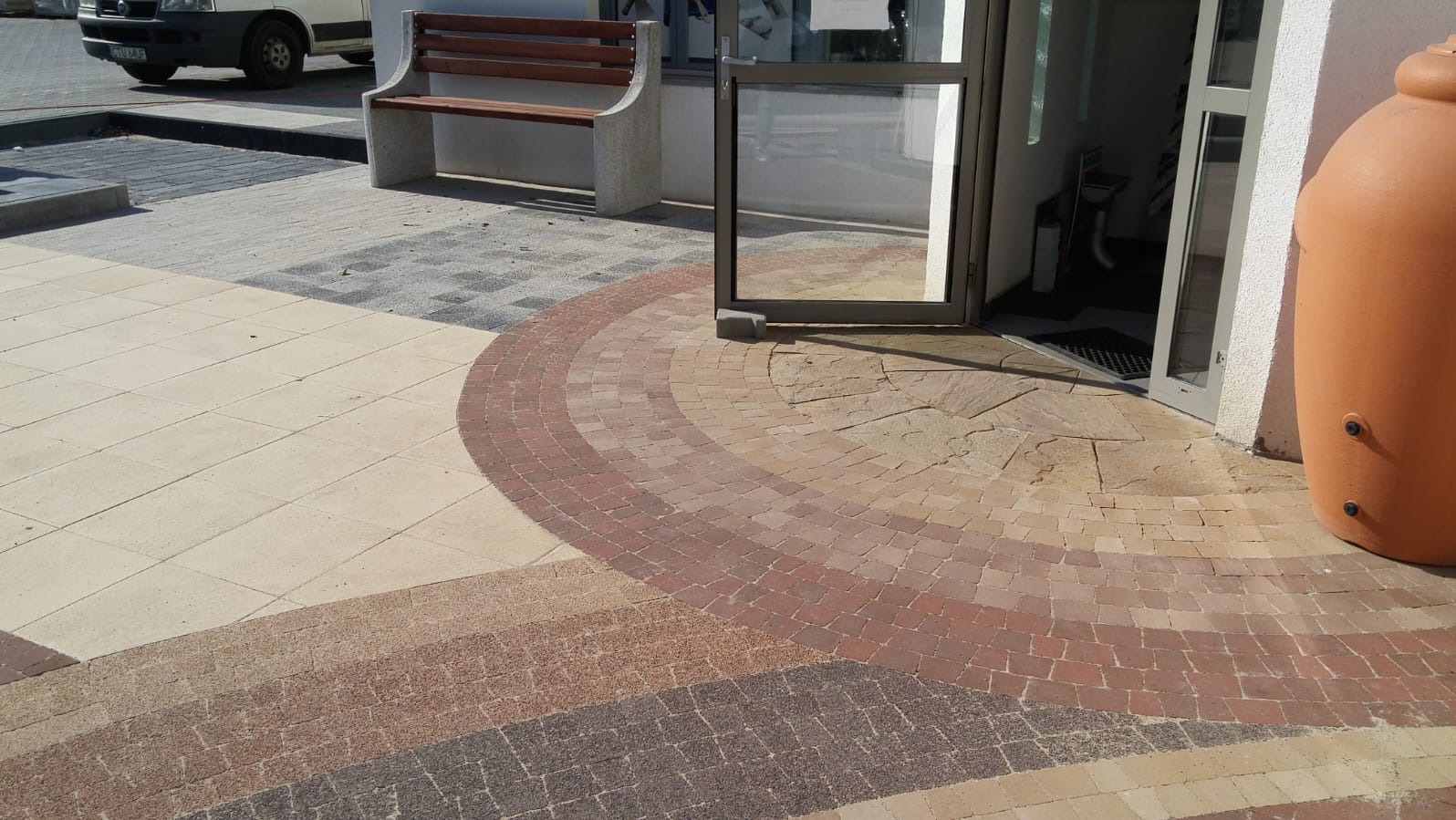 Kolorowa nawierzchnia z kostki brukowej ułożona w łukowate wzory przed wejściem do budynku. Widoczna ławka i duży, pomarańczowy zbiornik na wodę.