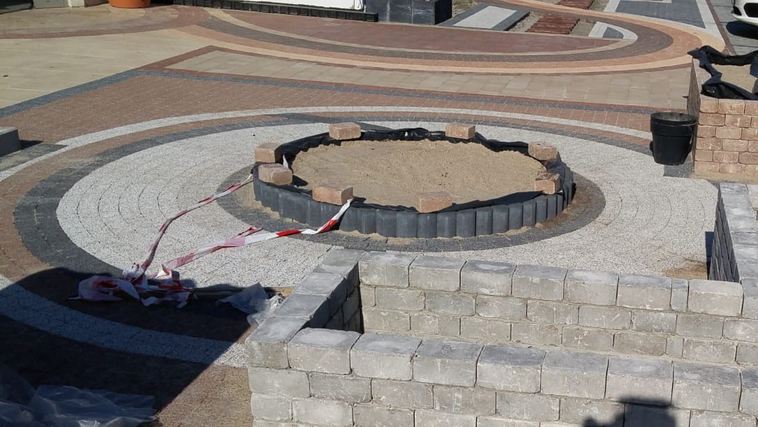 Okrągła rabata z piaskiem otoczona kostką, fragment murku z szarej kostki, wzorzysta nawierzchnia z kostki brukowej w odcieniach szarości i brązu, taśma ostrzegawcza.