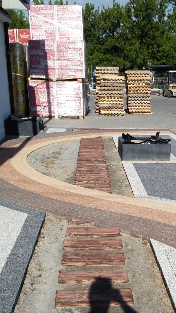 Aranżacja przestrzeni zewnętrznej z drewnopodobną ścieżką i różnymi rodzajami kostki brukowej. W tle palety z materiałami budowlanymi i wózki widłowe.