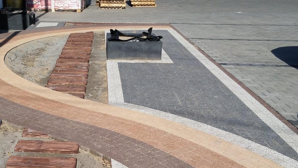 Aranżacja przestrzeni z kostki brukowej w odcieniach brązu i szarości, z elementami drewnopodobnymi i donicą. Widok na ścieżkę i plac z kostki.