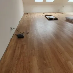 Montaż paneli podłogowych w poddaszu z oknami dachowymi; widoczny kabel i poziomica przy ścianie.