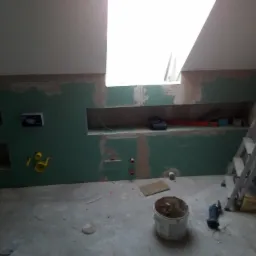 Wnętrze łazienki w trakcie prac wykończeniowych, z widocznymi płytami gipsowo-kartonowymi na ścianach, przygotowanymi podłoże pod armaturę i instalacje, oraz narzędziami pracy w pomieszczeniu.