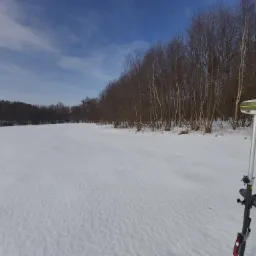 Pomiarowy odbiornik GNSS na tyczce, ustawiony na zasypanym śniegiem terenie, w tle linia drzew pod błękitnym, pochmurnym niebem.
