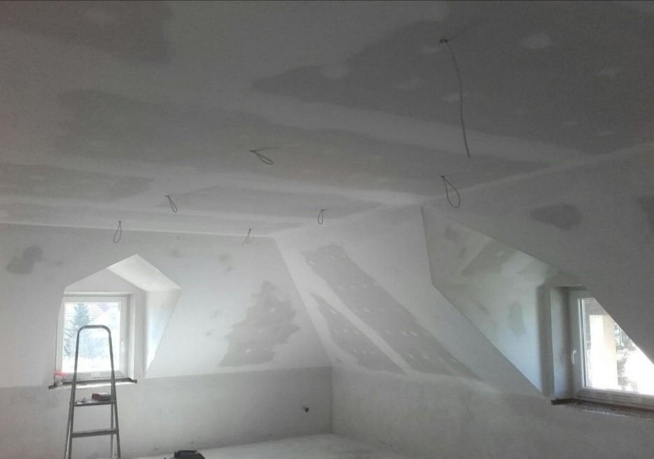 Poddasze w trakcie wykańczania: ściany i sufit pokryte gładzią gipsową, widoczne kable wystające z sufitu, okno dachowe z drabinką opartą o parapet.