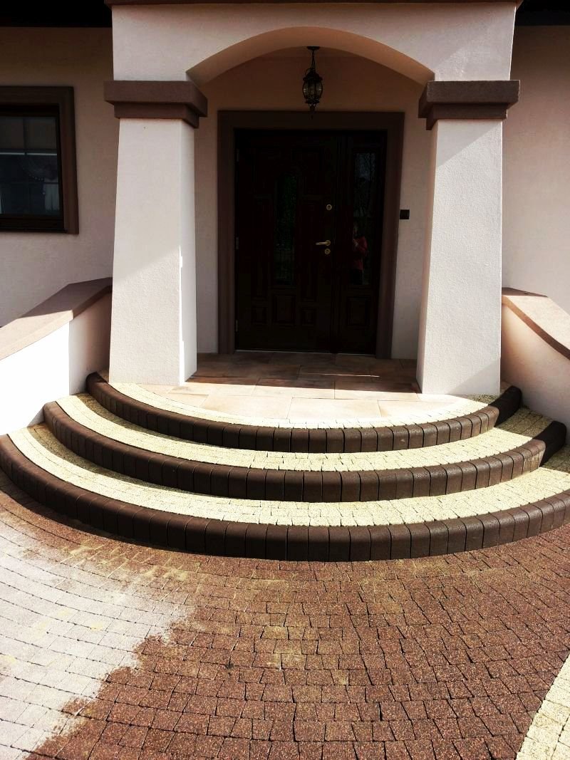 Półokrągłe schody wejściowe z dwukolorowej kostki, prowadzące do ciemnych drzwi wejściowych domu z kolumnami i łukiem.