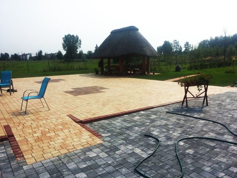 Ułożona nawierzchnia z kostki w dwóch odcieniach szarości i beżu, z czerwoną obwódką, altana z trzcinowym dachem w tle, dwa niebieskie krzesła i wąż ogrodowy na nowej nawierzchni.