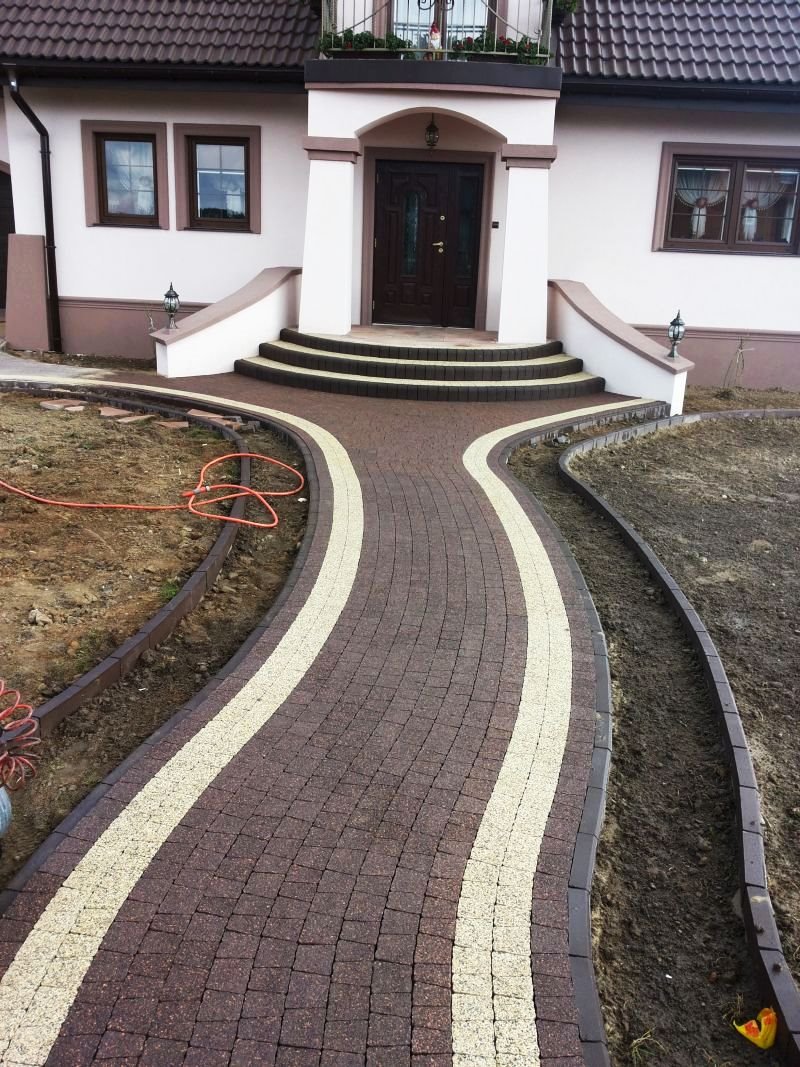 Brązowa ścieżka z jasnymi pasami, ułożona z kostki, prowadząca do wejścia do domu z brązowymi oknami i drzwiami, widoczny fragment trawnika i pomarańczowy wąż.