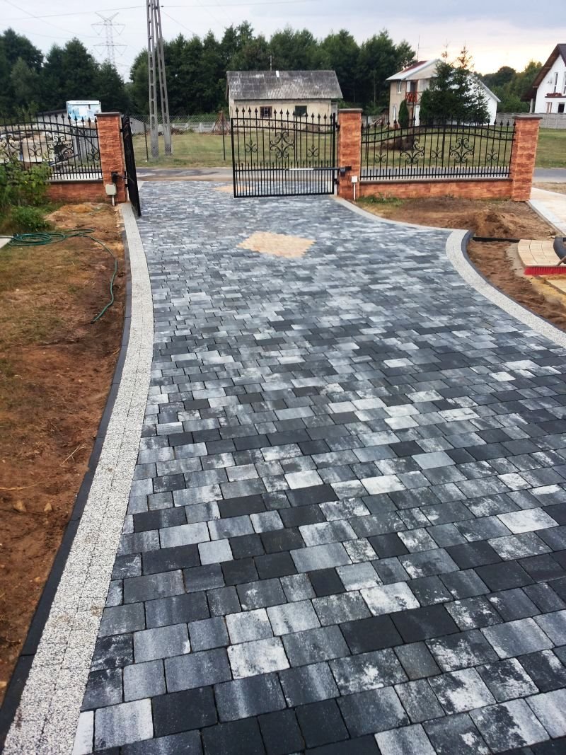 Szeroki podjazd z szarej, melanżowej kostki prowadzący do bramy z kutego żelaza na ceglanych słupkach, widok z góry na ukończoną realizację.