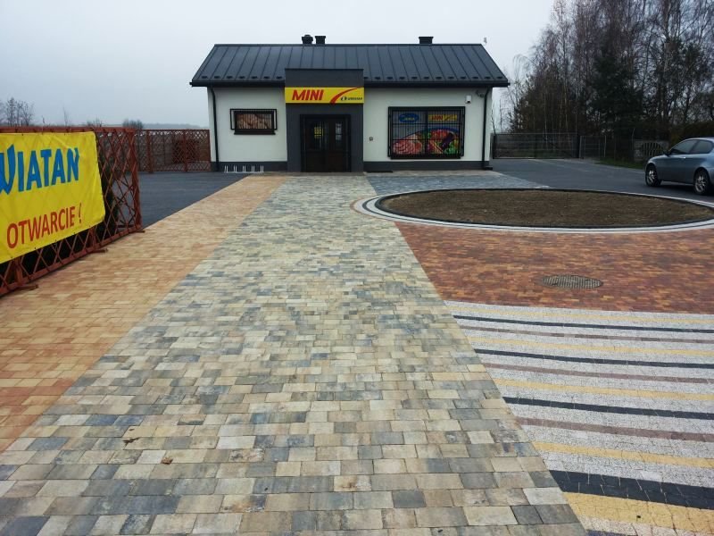 Nowo ułożona, wielobarwna nawierzchnia z kostki brukowej przed sklepem Mini, z okrągłym klombem po prawej i banerem reklamowym po lewej stronie, widok z perspektywy pieszego.
