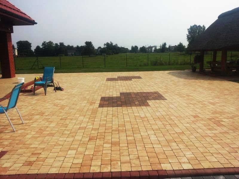 Ułożona kostka brukowa w odcieniach beżu i brązu tworząca wzór na podwórku z altaną krytą strzechą w tle, widoczne dwa niebieskie krzesła ogrodowe.