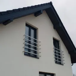 Fragment ściany szczytowej domu w stanie deweloperskim z oknami zabezpieczonymi metalowymi barierkami i ciemnym dachem z rynną