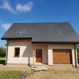Niedawno ukończony, parterowy dom jednorodzinny z szarą dachówką, drewnianą bramą garażową i oknami dachowymi, na działce w trakcie zagospodarowywania.