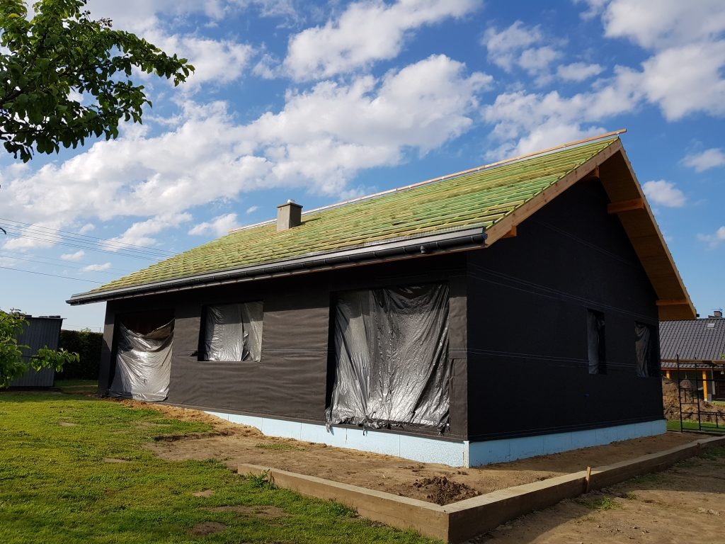 Budowa domu jednorodzinnego w Łaziskach: widok na budynek z czarną membraną na ścianach, zabezpieczone okna folią, zielony dach w trakcie budowy i fundamenty z niebieskim styropianem.
