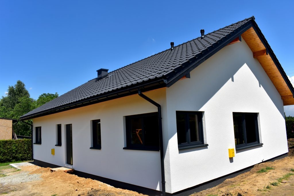 Nowoczesny, parterowy dom z ciemną dachówką i białymi ścianami zewnętrznymi, widoczne elementy wykończeniowe dachu i czarne okna, teren wokół domu w trakcie prac ziemnych.