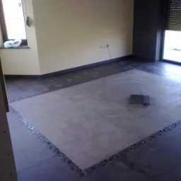 Układanie płytek podłogowych w pomieszczeniu, widoczne fragmenty nowej i starej podłogi, płytki ułożone w centralnej części z ozdobnym pasem mozaiki na obrzeżach, w tle ściany i okno.