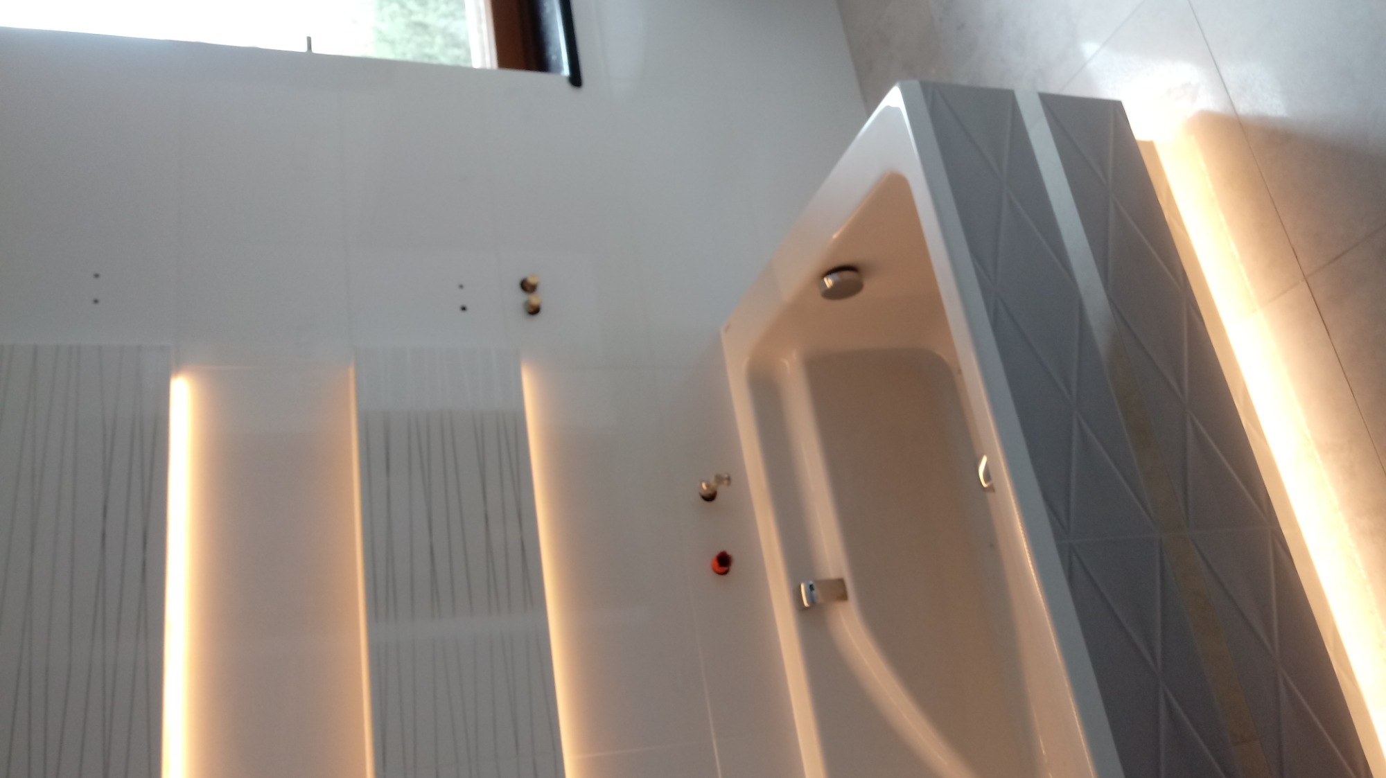 Nowoczesna biała wanna z geometrycznym wzorem na obudowie, podświetlona listwami LED, zamontowana w minimalistycznej łazience z białymi płytkami i ukrytymi przyłączami.