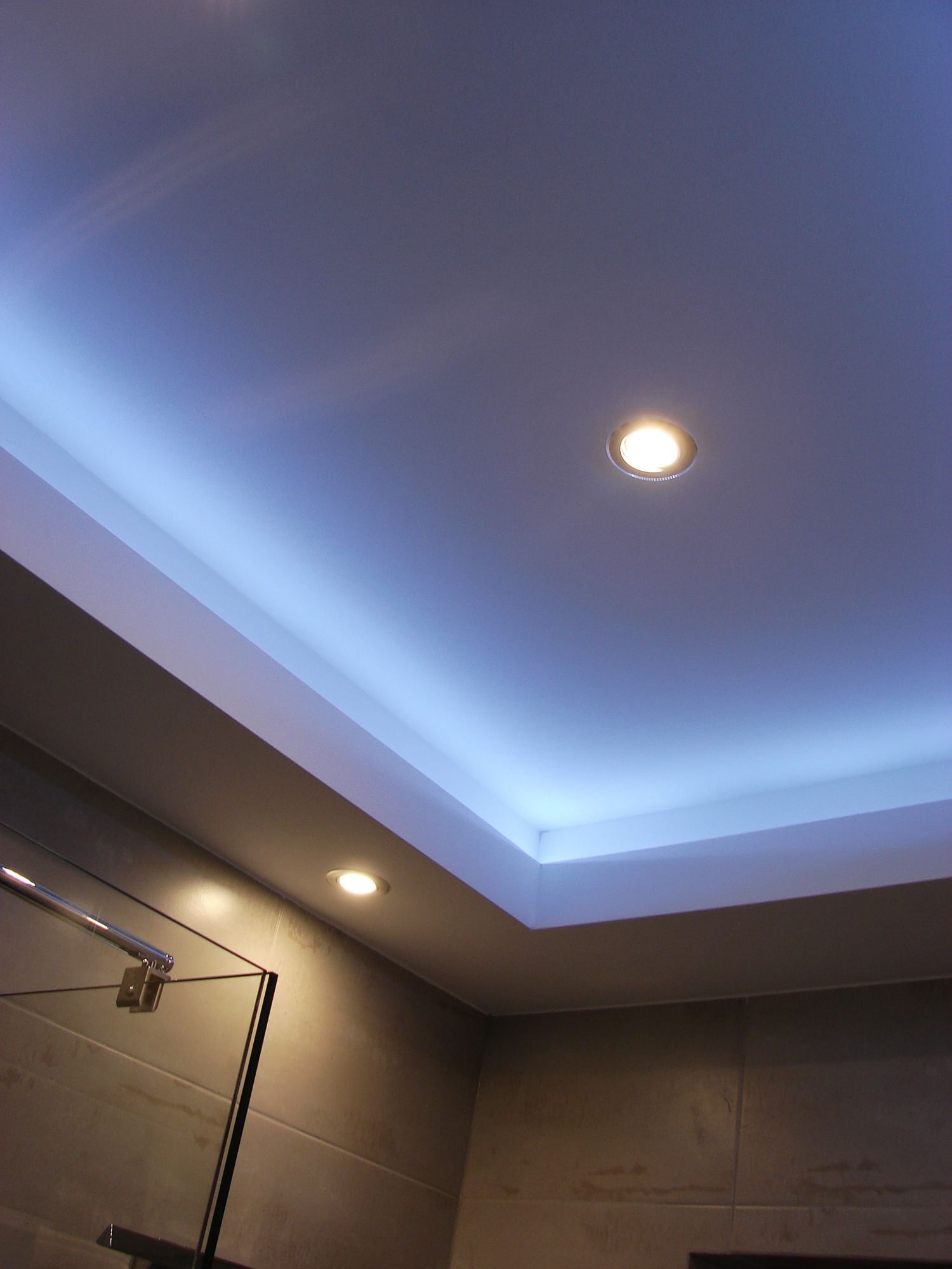 Podwieszany sufit w łazience z oświetleniem LED w kolorze niebieskim, widoczne dwie okrągłe lampy halogenowe, fragment kabiny prysznicowej z chromowanymi elementami.