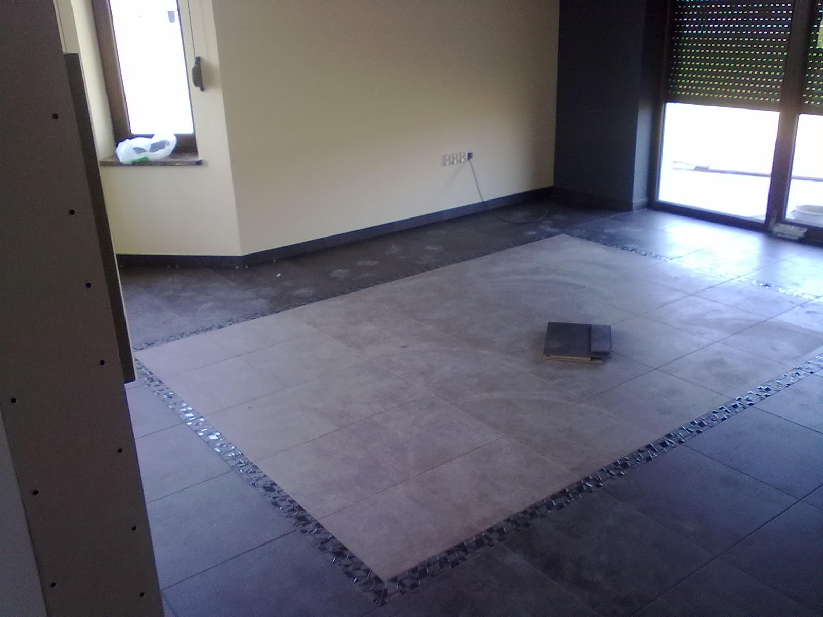 Układanie płytek podłogowych w pomieszczeniu, widoczne fragmenty nowej i starej podłogi, płytki ułożone w centralnej części z ozdobnym pasem mozaiki na obrzeżach, w tle ściany i okno.