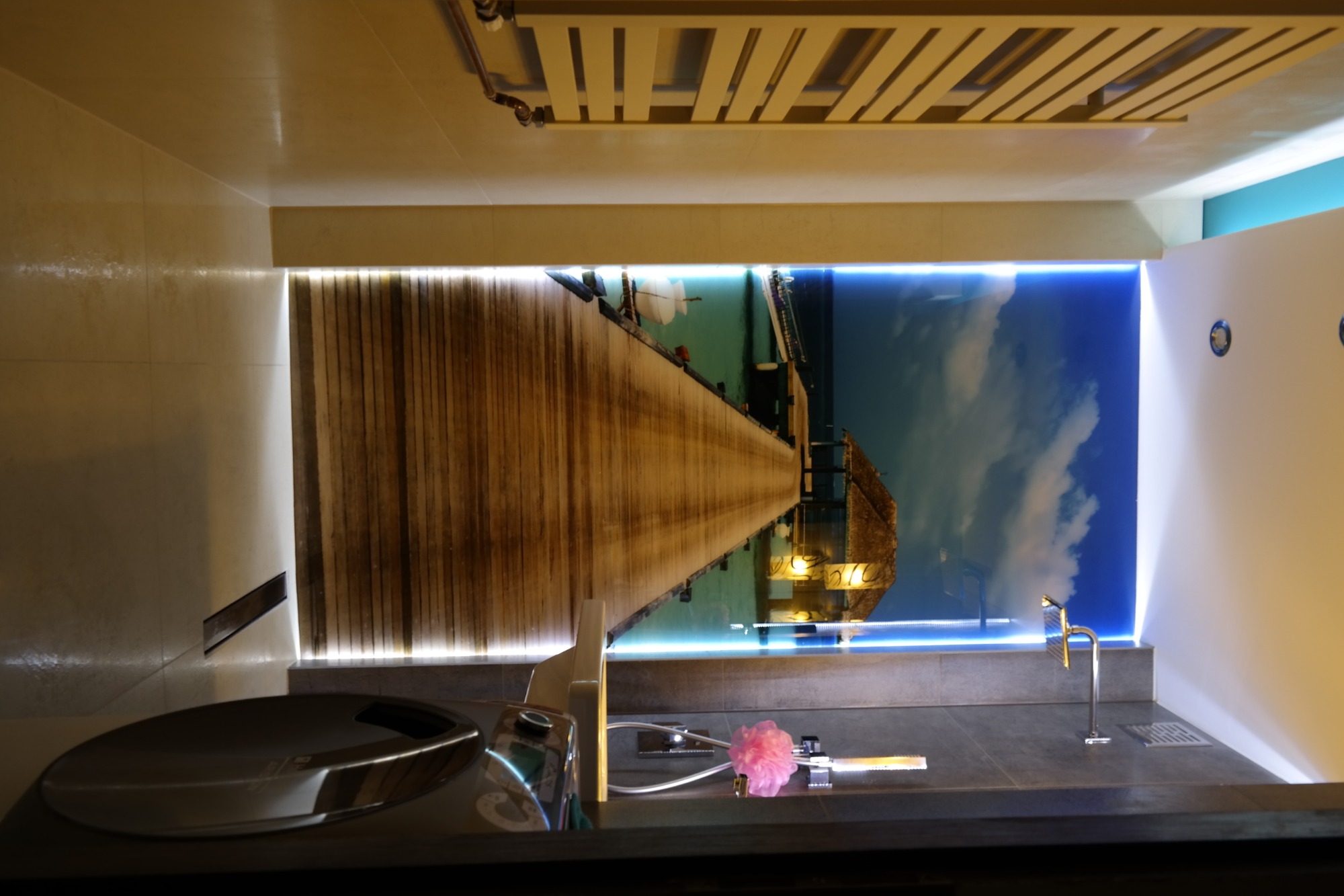 Nowoczesna łazienka z podświetlanym panelem ściennym przedstawiającym drewniany pomost nad wodą, szara umywalka nablatowa i chromowana armatura.