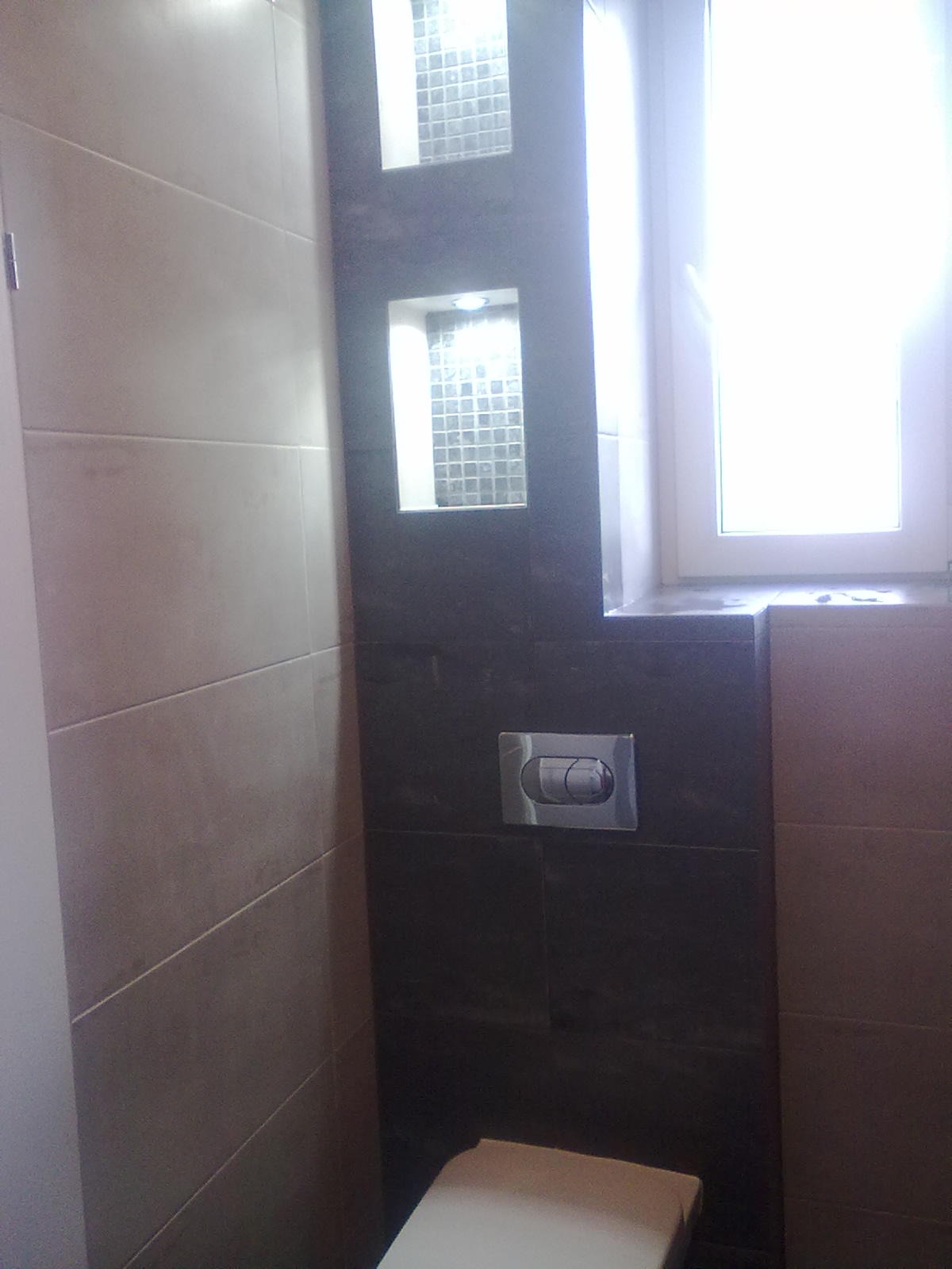 Wykończona łazienka z beżowymi i ciemnymi płytkami, wnęki z mozaiką oraz przycisk spłuczki na ciemnej ścianie, widok z dołu na toaletę.