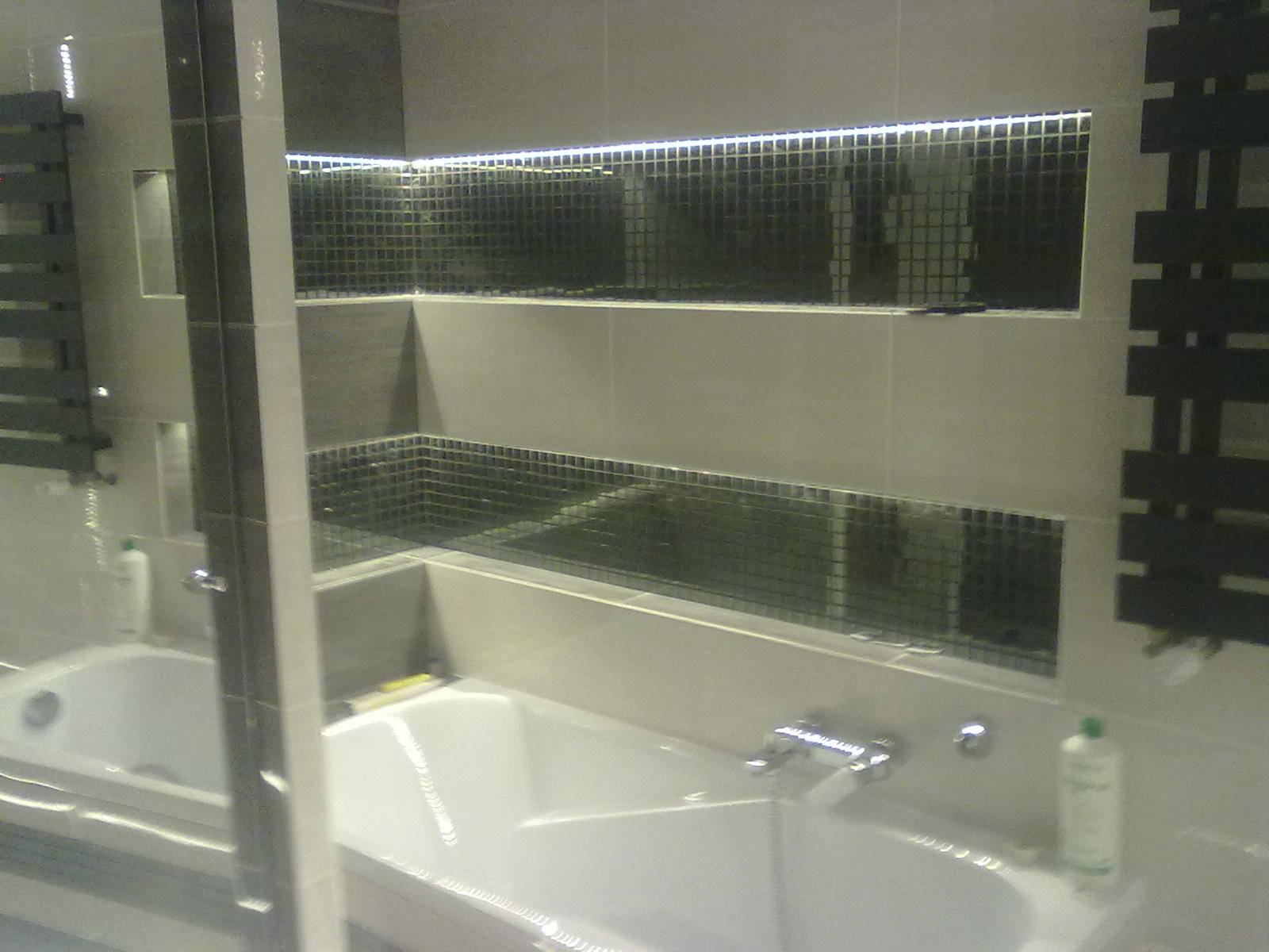 Nowoczesna łazienka z wanną, mozaiką w niszach podświetlonych LED i grzejnikiem drabinkowym, widziana przez szklaną ściankę prysznicową.