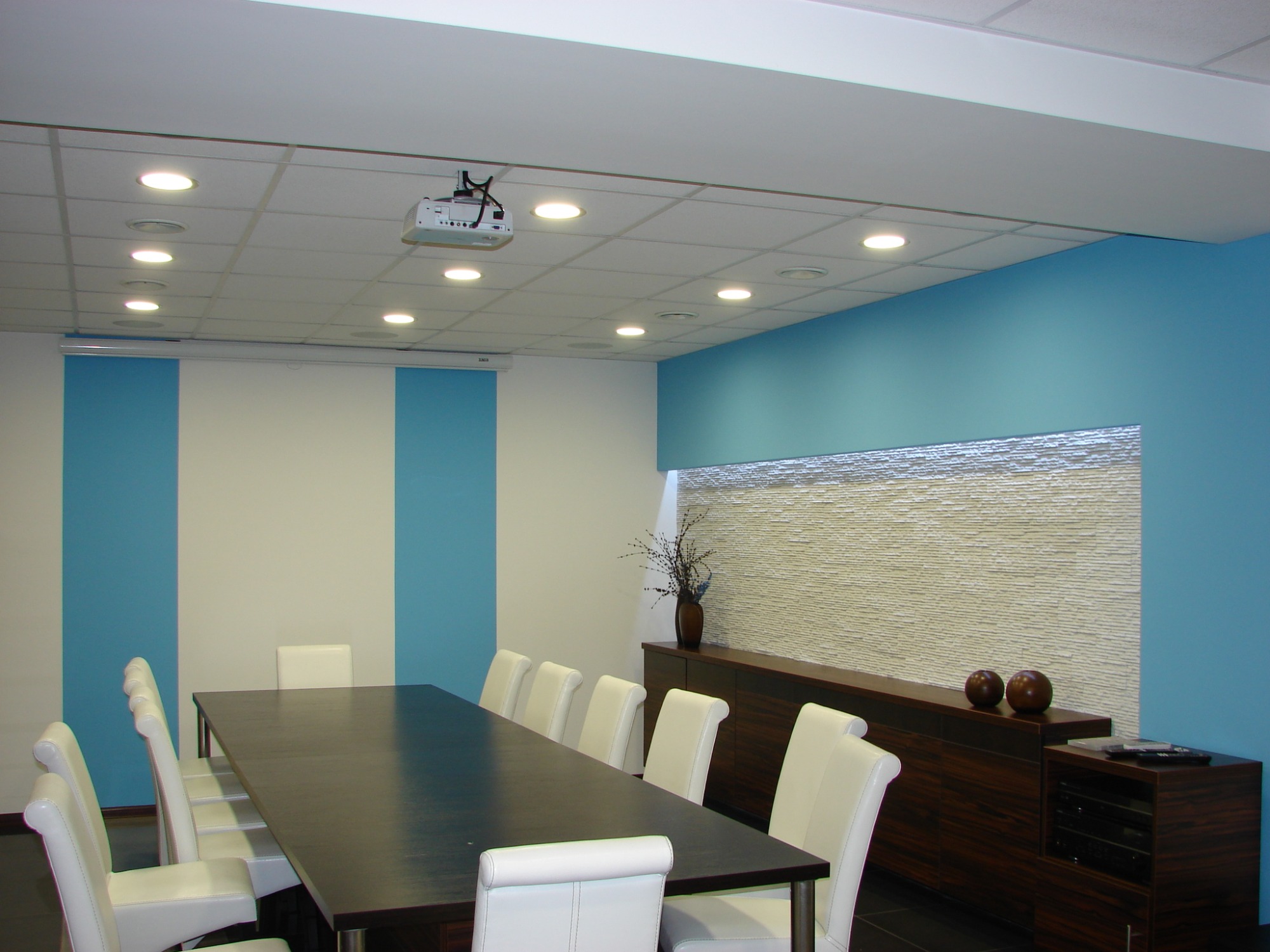 Nowoczesna sala konferencyjna z długim, ciemnym stołem, białymi krzesłami, niebieskimi akcentami na ścianach i dekoracyjną ścianą z kamienia za ciemnym kredensem.