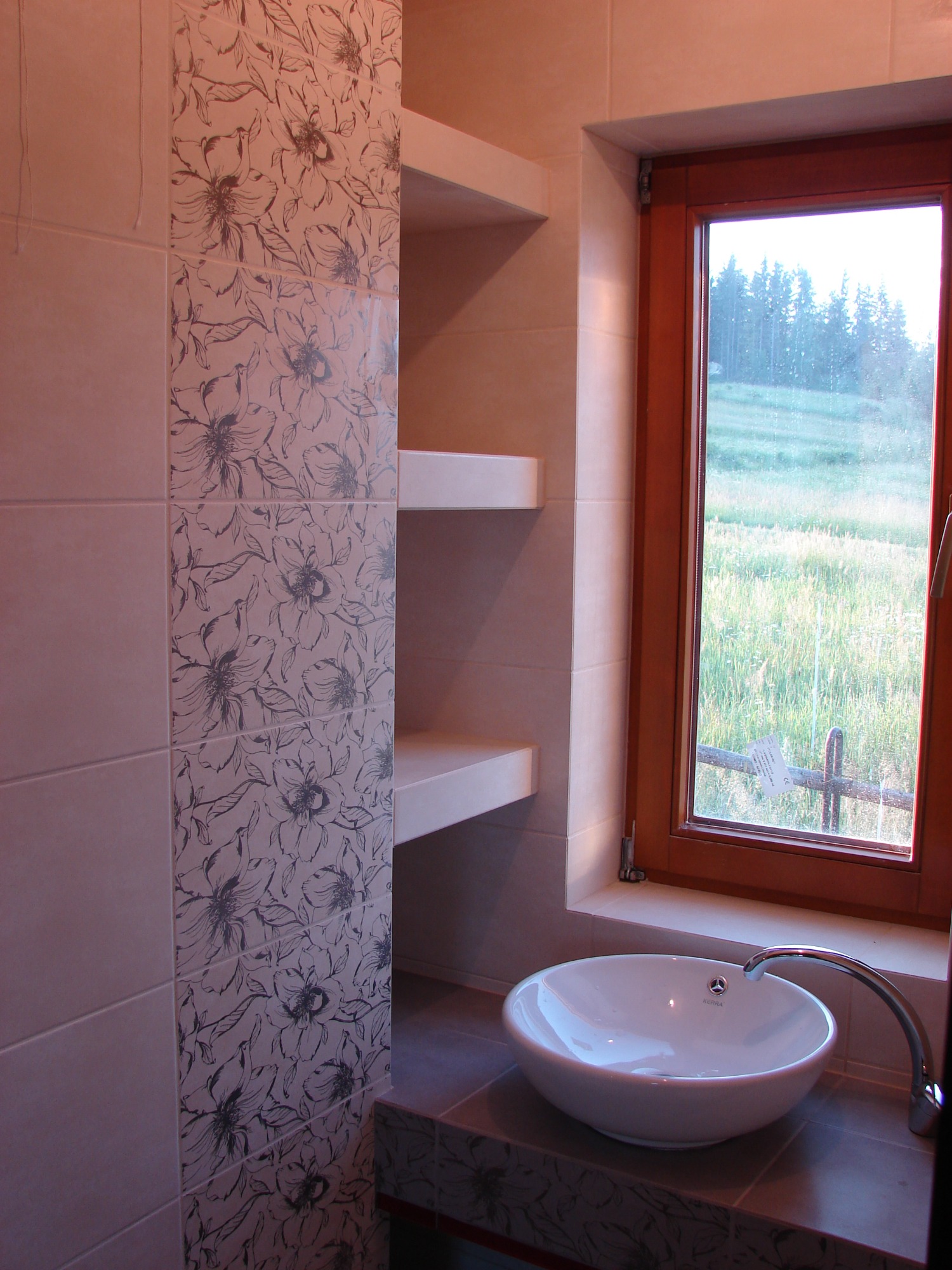 Jasna łazienka z umywalką nablatową, płytkami z motywem kwiatowym i widokiem na zieleń za oknem.