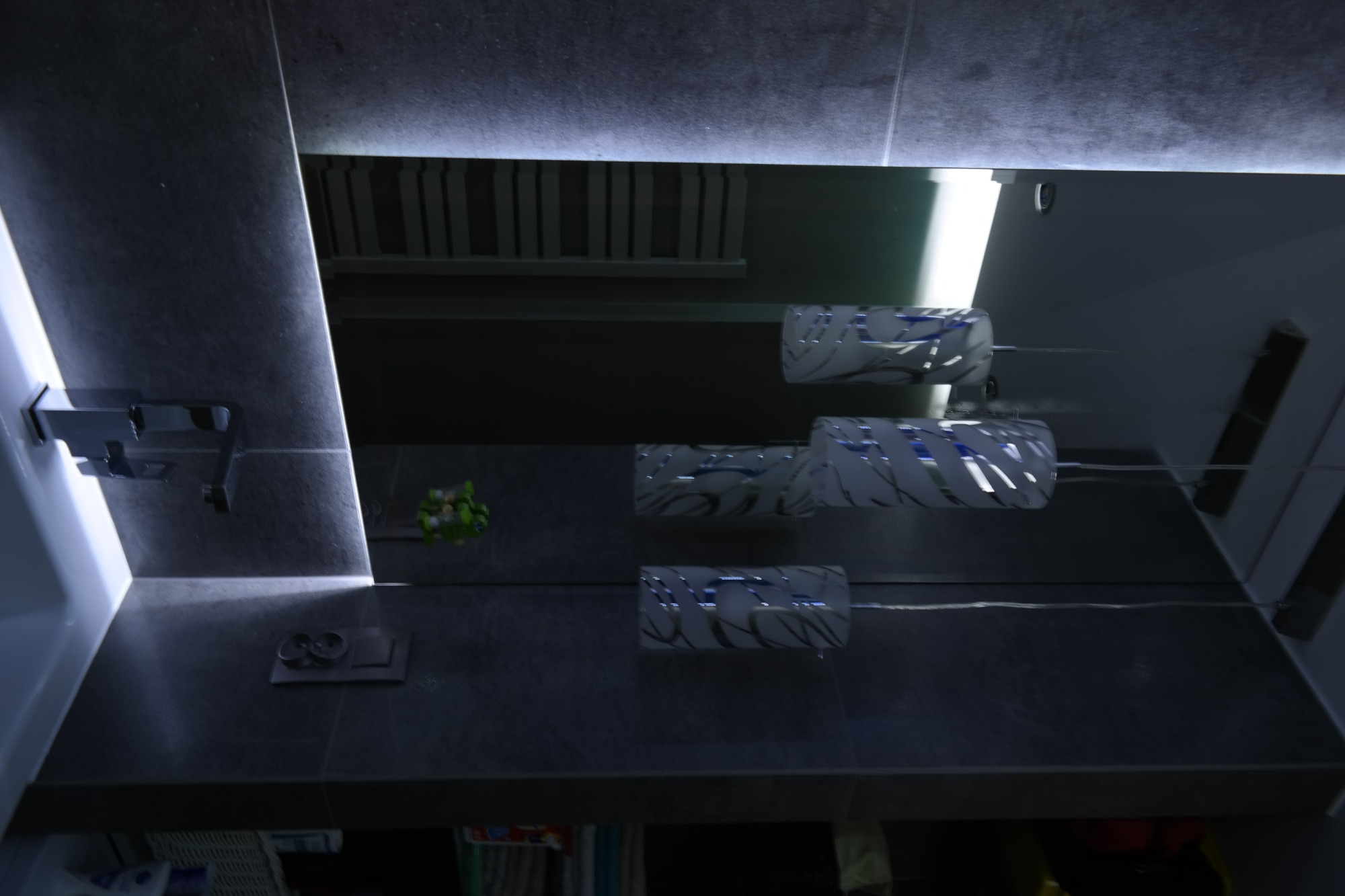 Szara łazienka z trzema nowoczesnymi lampami ściennymi z białymi abażurami i wzorem w liście, ciemne płytki, bateria umywalkowa i akcesoria łazienkowe.