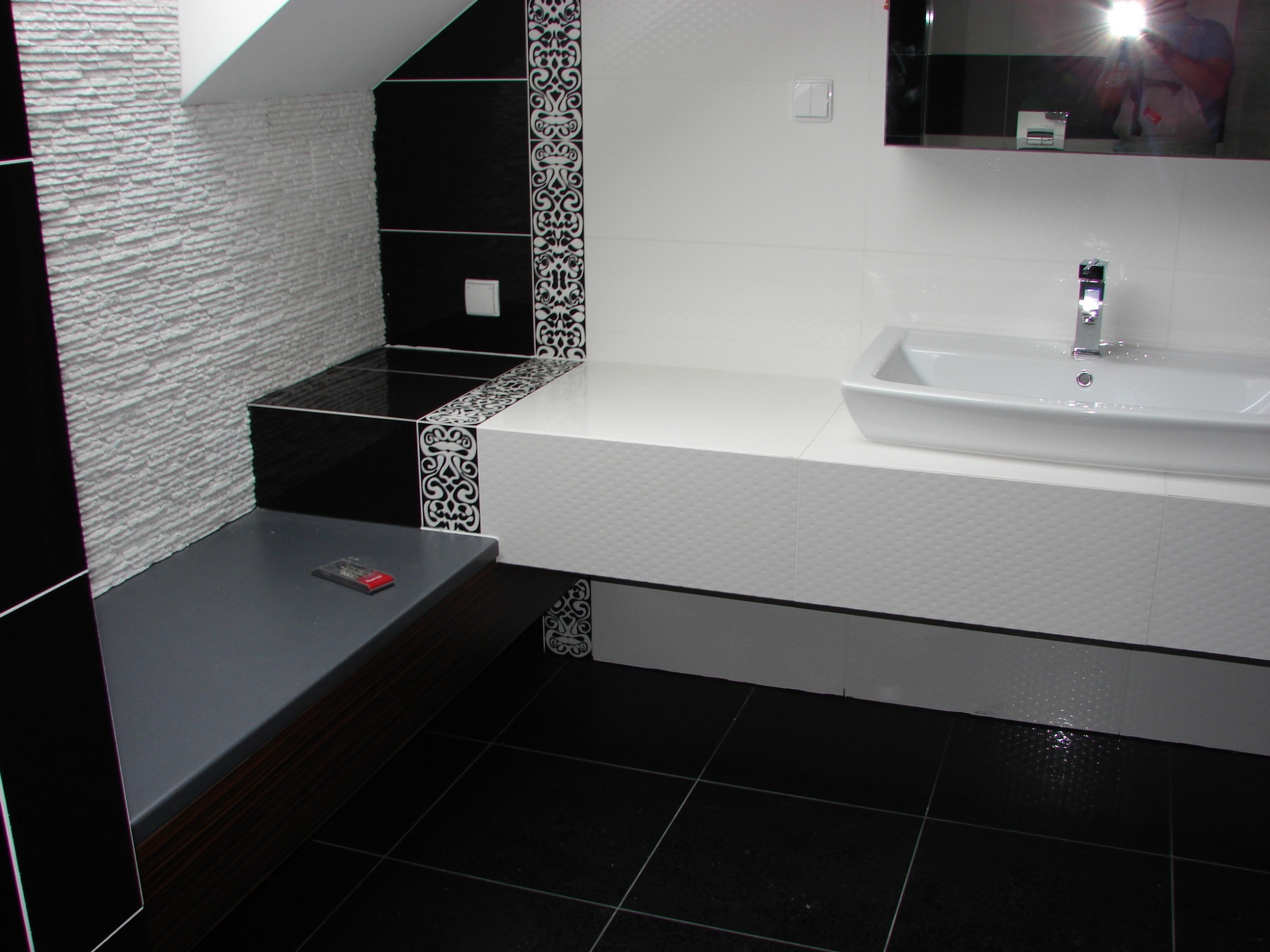 Nowoczesna łazienka w czarno-białej kolorystyce z umywalką nablatową, dekoracyjną mozaiką i strukturalną ścianą, widoczny pilot na półce.