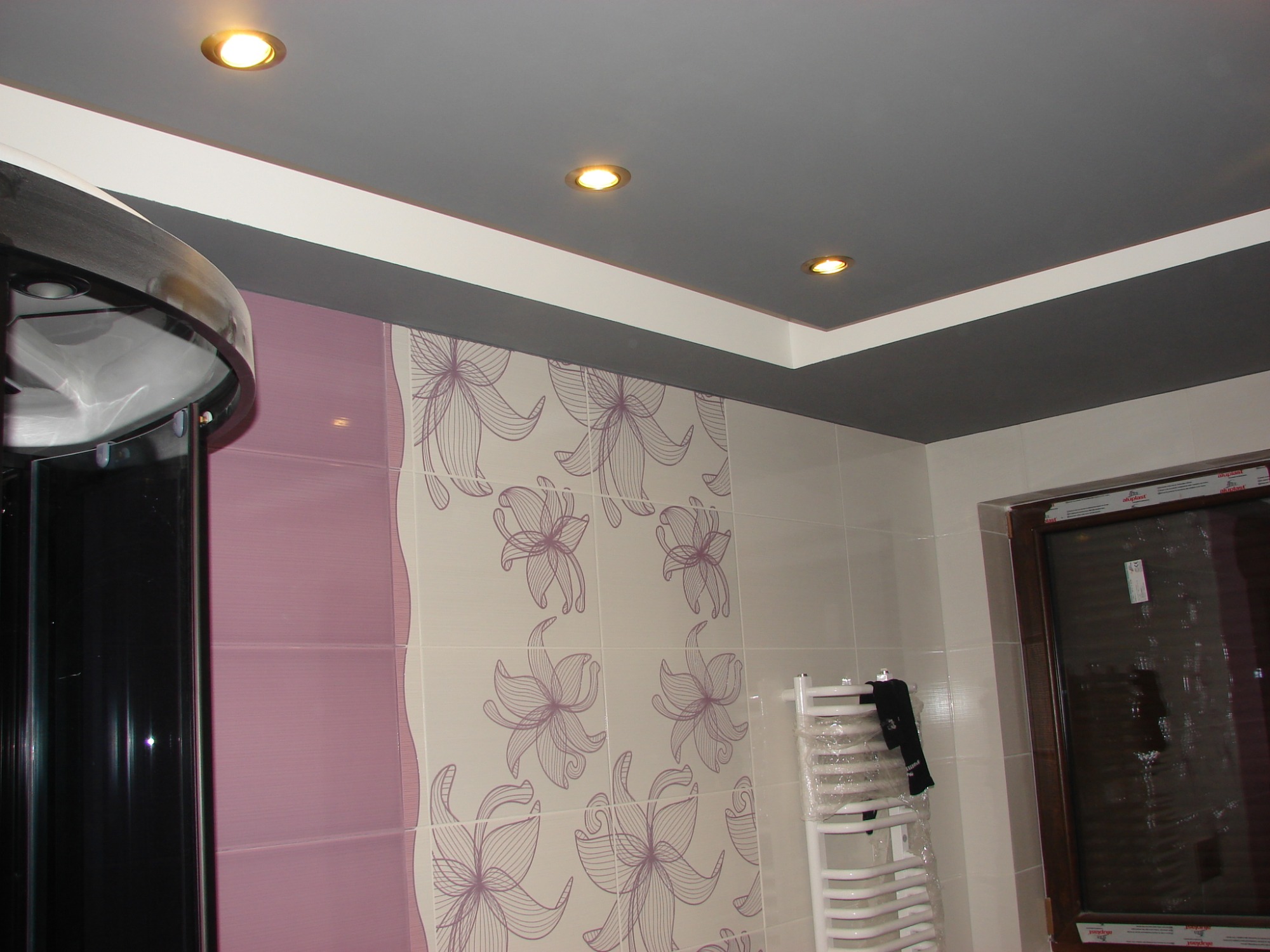 Wykończona łazienka z szarą, podwieszaną częścią sufitu z trzema halogenami, ścianami wyłożonymi płytkami w odcieniach fioletu i bieli z motywem kwiatowym oraz grzejnikiem drabinkowym owiniętym...