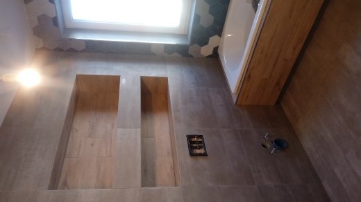 Łazienka w trakcie wykańczania: widoczne wnęki w ścianie wyłożone płytkami imitującymi drewno, heksagonalne płytki pod oknem, obudowa wanny z drewnopodobnych paneli, puszka elektryczna i rury...