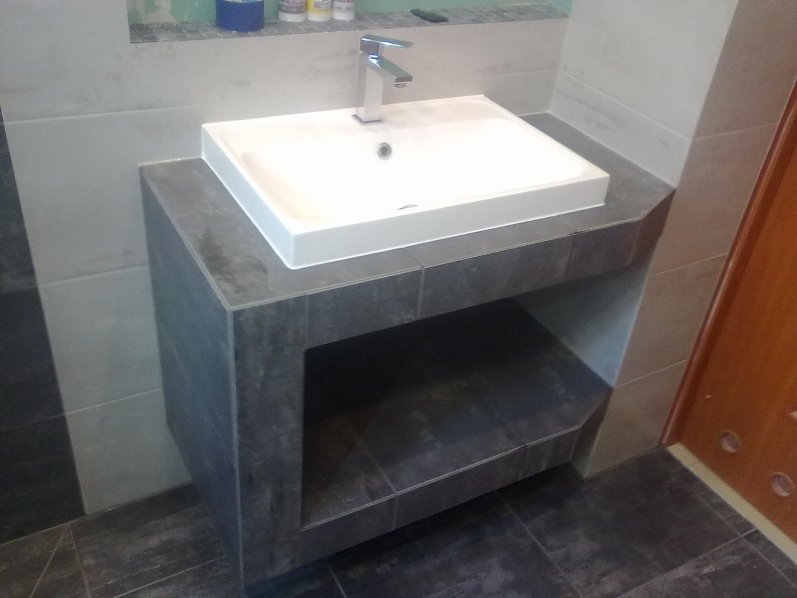 Nowoczesna umywalka nablatowa z chromowaną baterią, osadzona na szafce z szarych płytek, z otwartą półką, w łazience wykończonej płytkami w odcieniach szarości.