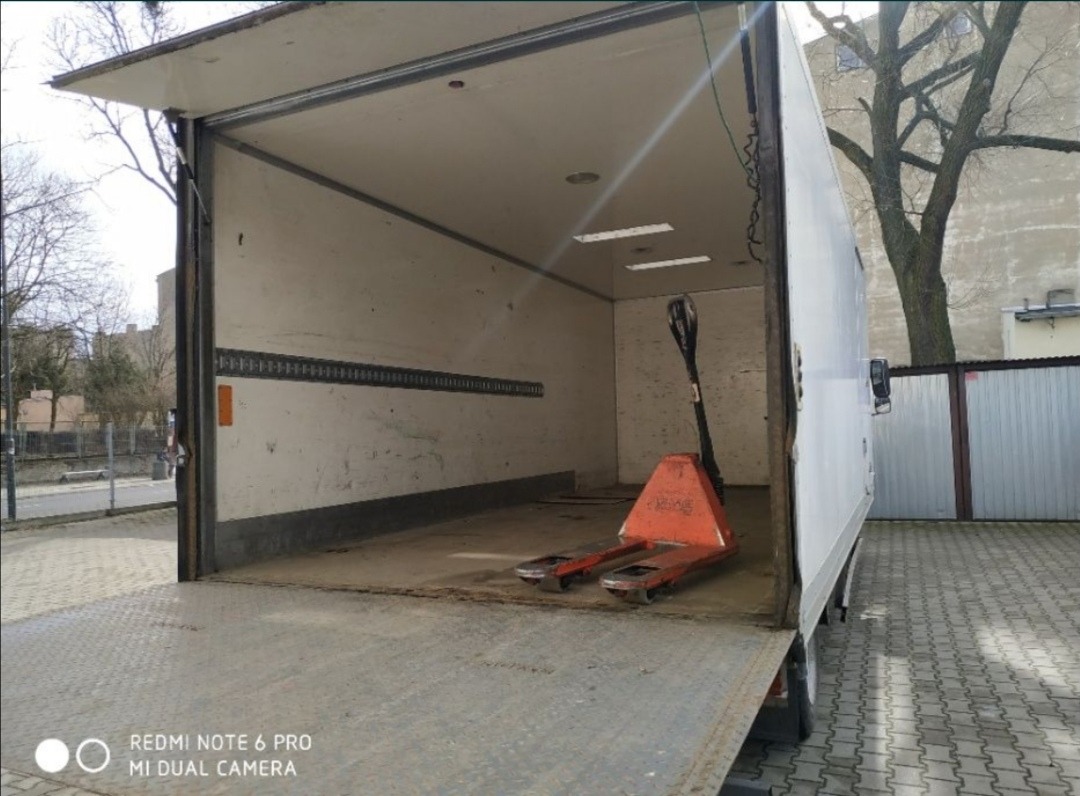 Wnętrze paki białego samochodu dostawczego z otwartą klapą załadunkową i wózkiem paletowym w środku, widok na ulicę i budynek w tle.