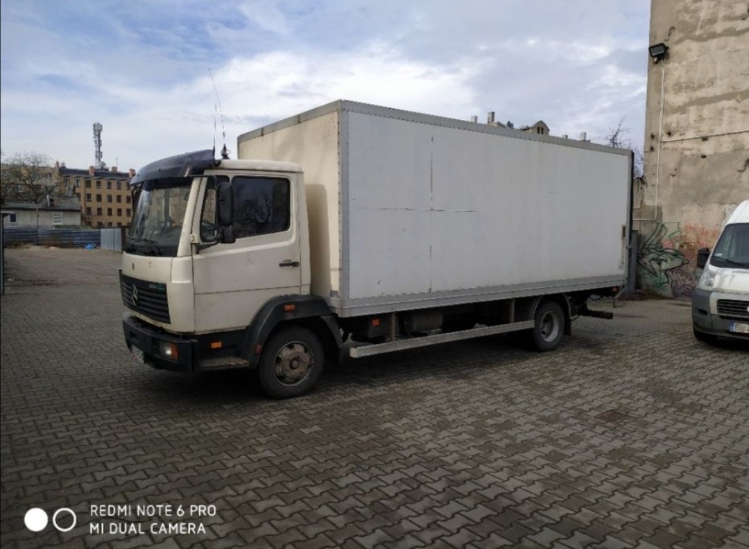 Biały samochód ciężarowy Mercedes-Benz z zabudową kontenerową, zaparkowany na brukowanym placu miejskim, widoczny w pochmurny dzień.