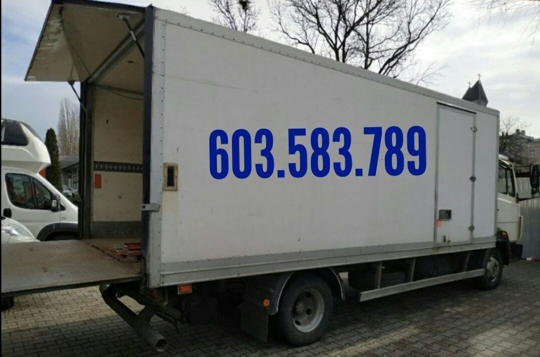 Biała ciężarówka z otwartą przestrzenią ładunkową i niebieskim numerem telefonu 603.583.789 na boku, zaparkowana na zewnątrz w pochmurny dzień.