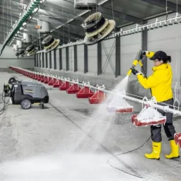 Pracownik w żółtej kurtce i kaloszach czyści przemysłową myjką ciśnieniową podwieszane karmniki w hali produkcyjnej.