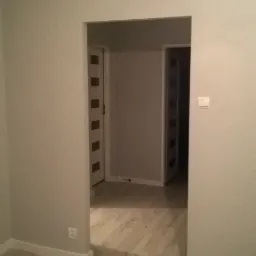 Wnętrze po remoncie: szare ściany, panele podłogowe imitujące drewno, widoczne dwie białe drzwi z przeszkleniami w głębi pomieszczenia.