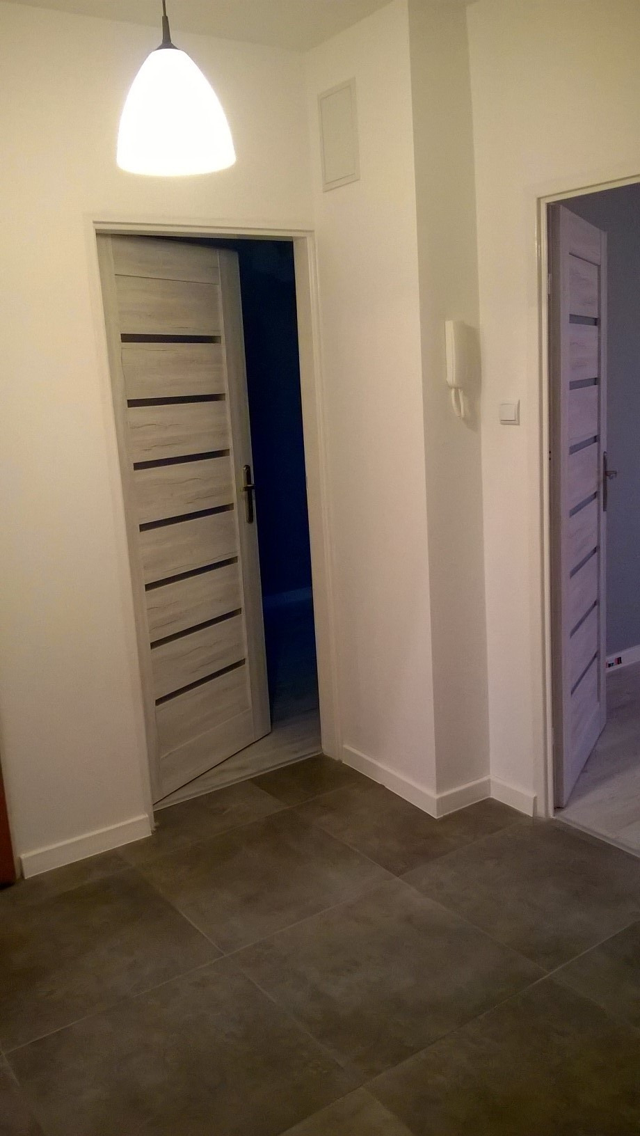 Wnętrze mieszkania po remoncie, widoczne dwie otwarte, nowoczesne drzwi w kolorze jasnego drewna z ciemnymi, poziomymi paskami, brązowe płytki podłogowe i białe ściany z widocznym domofonem.