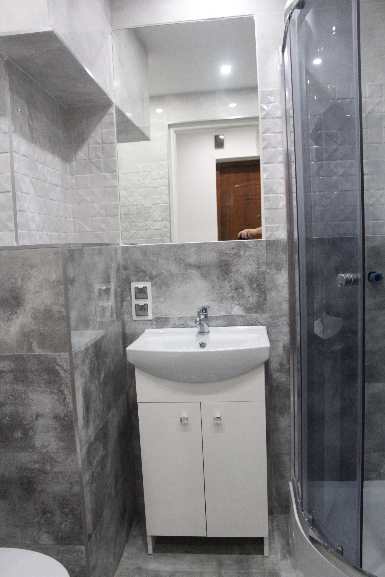 Nowoczesna łazienka z szarymi płytkami imitującymi beton, białą szafką pod umywalką z chromowanymi uchwytami, lustrem i kabiną prysznicową z transparentnego szkła.