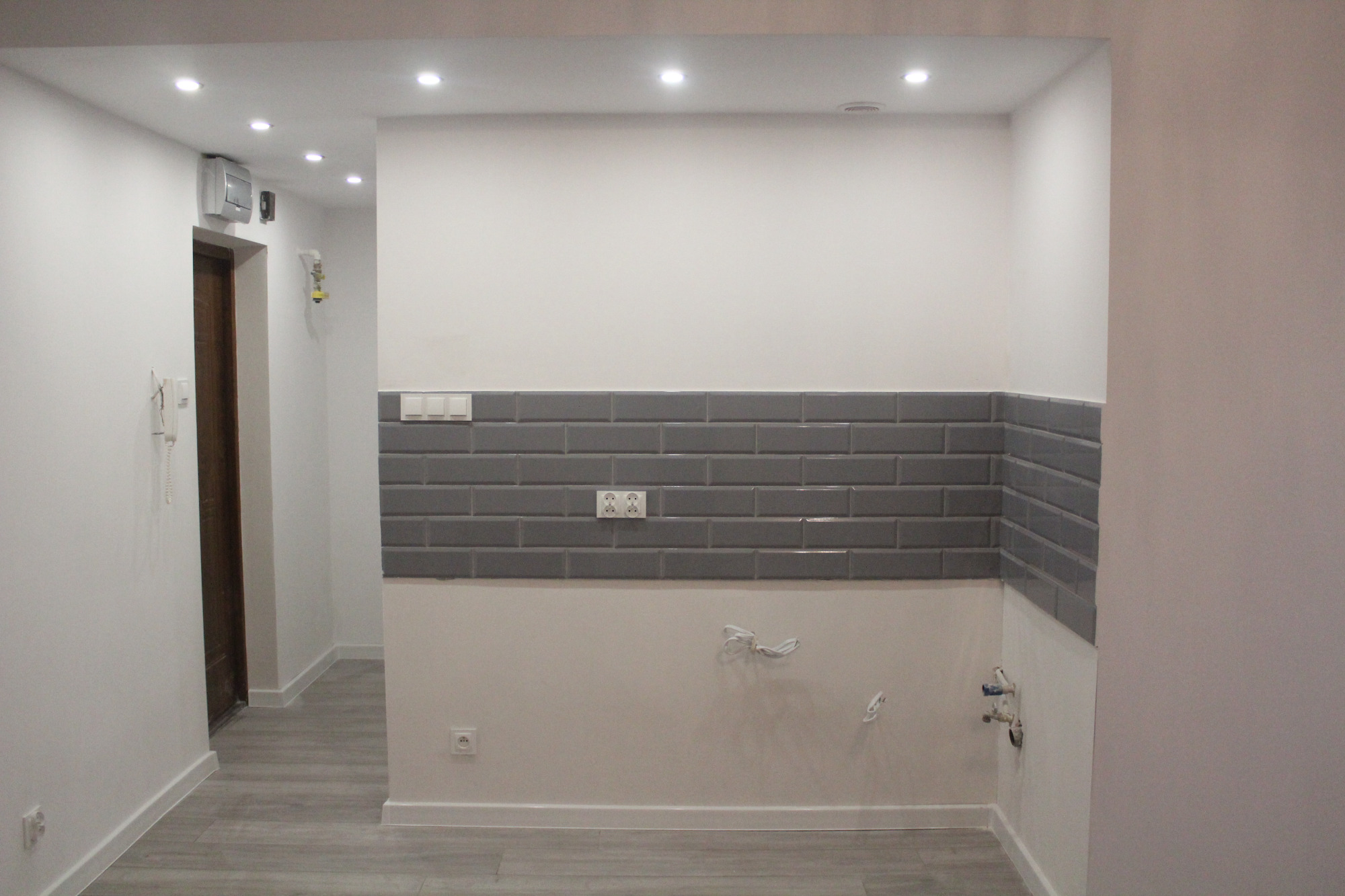 Świeżo wykafelkowana ściana szarymi, błyszczącymi płytkami w kuchni, widoczne gniazdka elektryczne i wyprowadzone rury wodne, przygotowane do dalszych prac wykończeniowych.