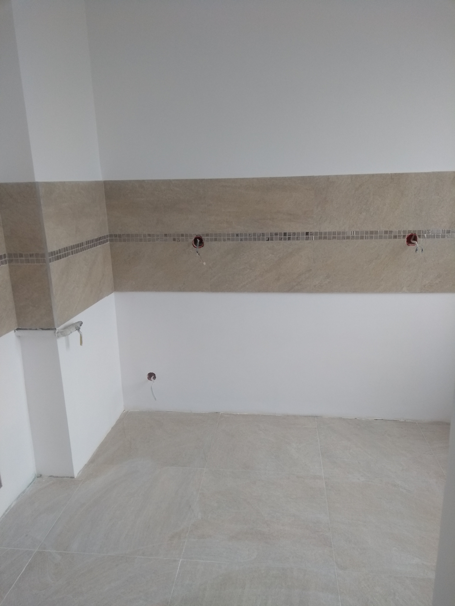 Wnętrze łazienki w trakcie remontu, widoczne położone płytki podłogowe i częściowo ułożona glazura z dekoracyjnym pasem mozaiki, wystające kable elektryczne ze ścian.