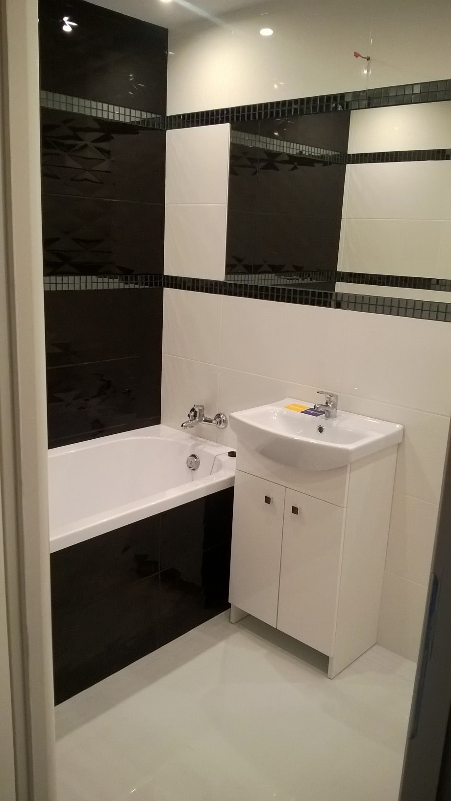Wykończona łazienka w czarno-białej kolorystyce z wanną i umywalką z szafką, ozdobiona mozaiką szklaną między płytkami.
