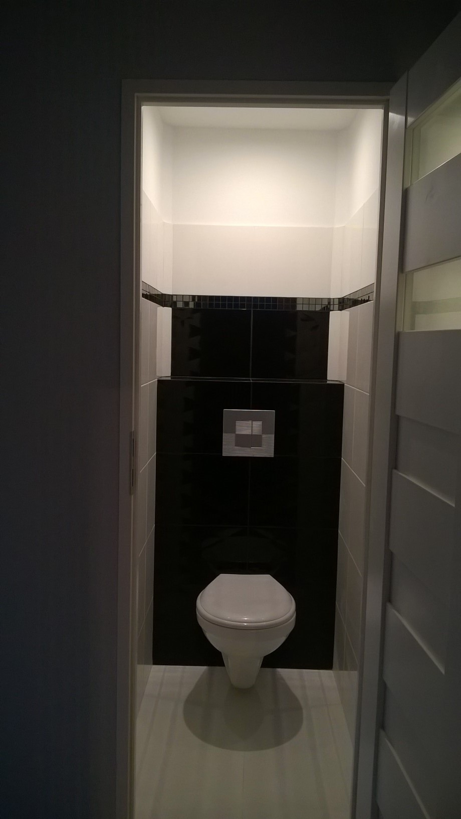 Wąska toaleta z podwieszaną muszlą klozetową, czarnymi płytkami do połowy ściany, wykończona mozaiką, z białymi płytkami powyżej i na podłodze, widziana z perspektywy wejścia.