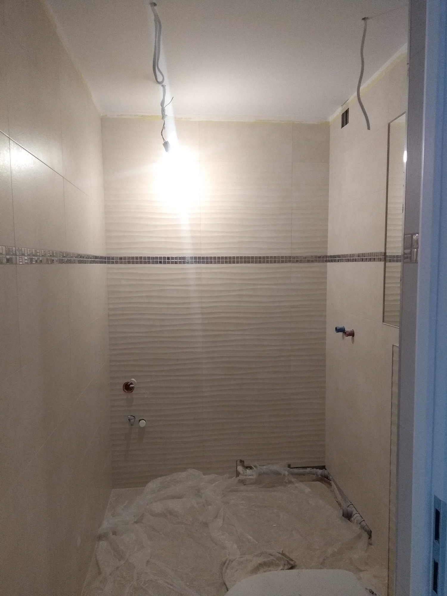 Łazienka w trakcie remontu, ściany wyłożone jasnymi płytkami z dekoracyjnym pasem mozaiki, podłoga zabezpieczona folią malarską, widoczne wystające przewody elektryczne i rury hydrauliczne.