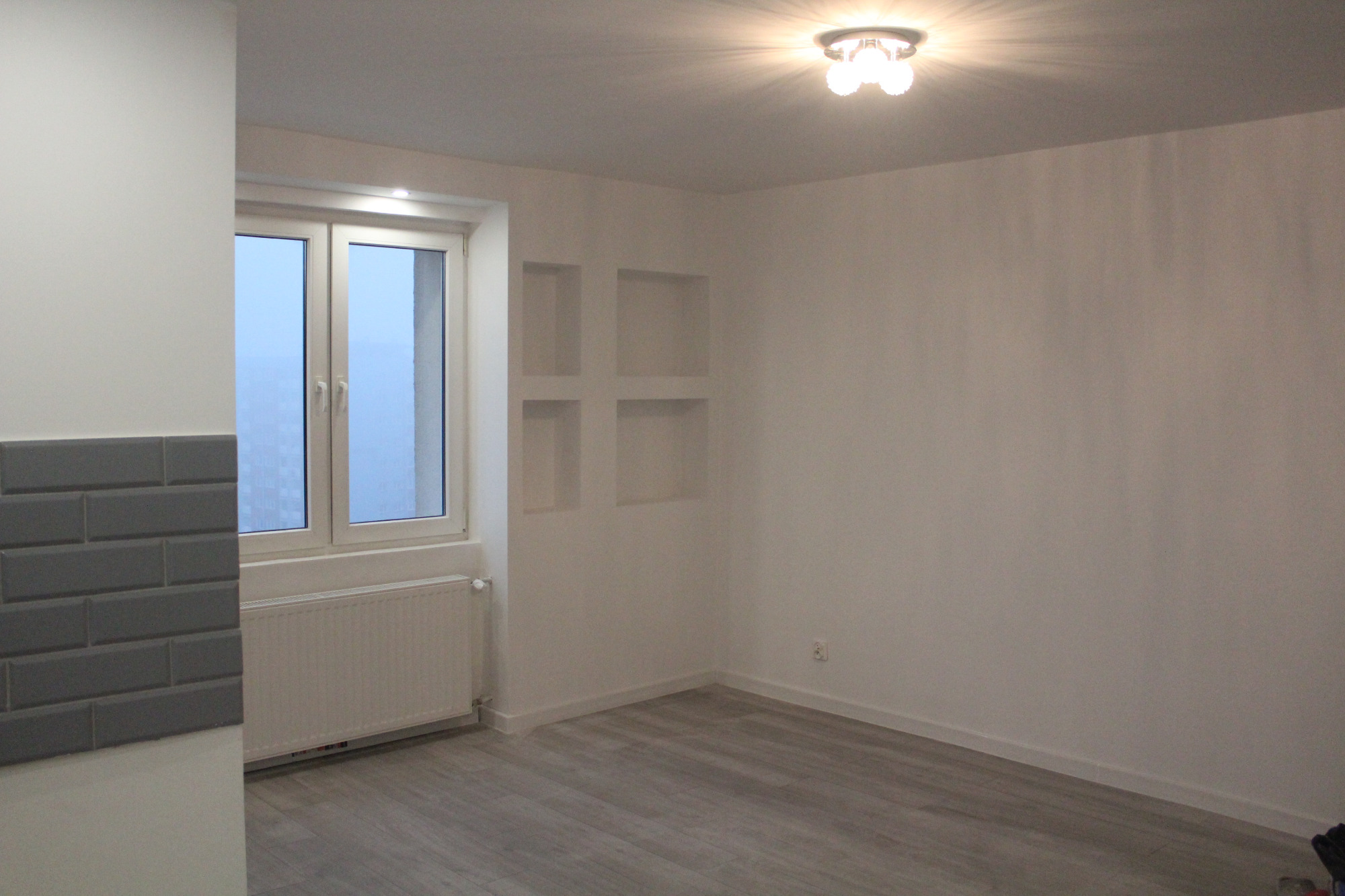 Pusty pokój po remoncie z szarą podłogą panelową, białymi ścianami z wnękami, oknem z grzejnikiem i fragmentem ściany z szarymi płytkami typu metro.